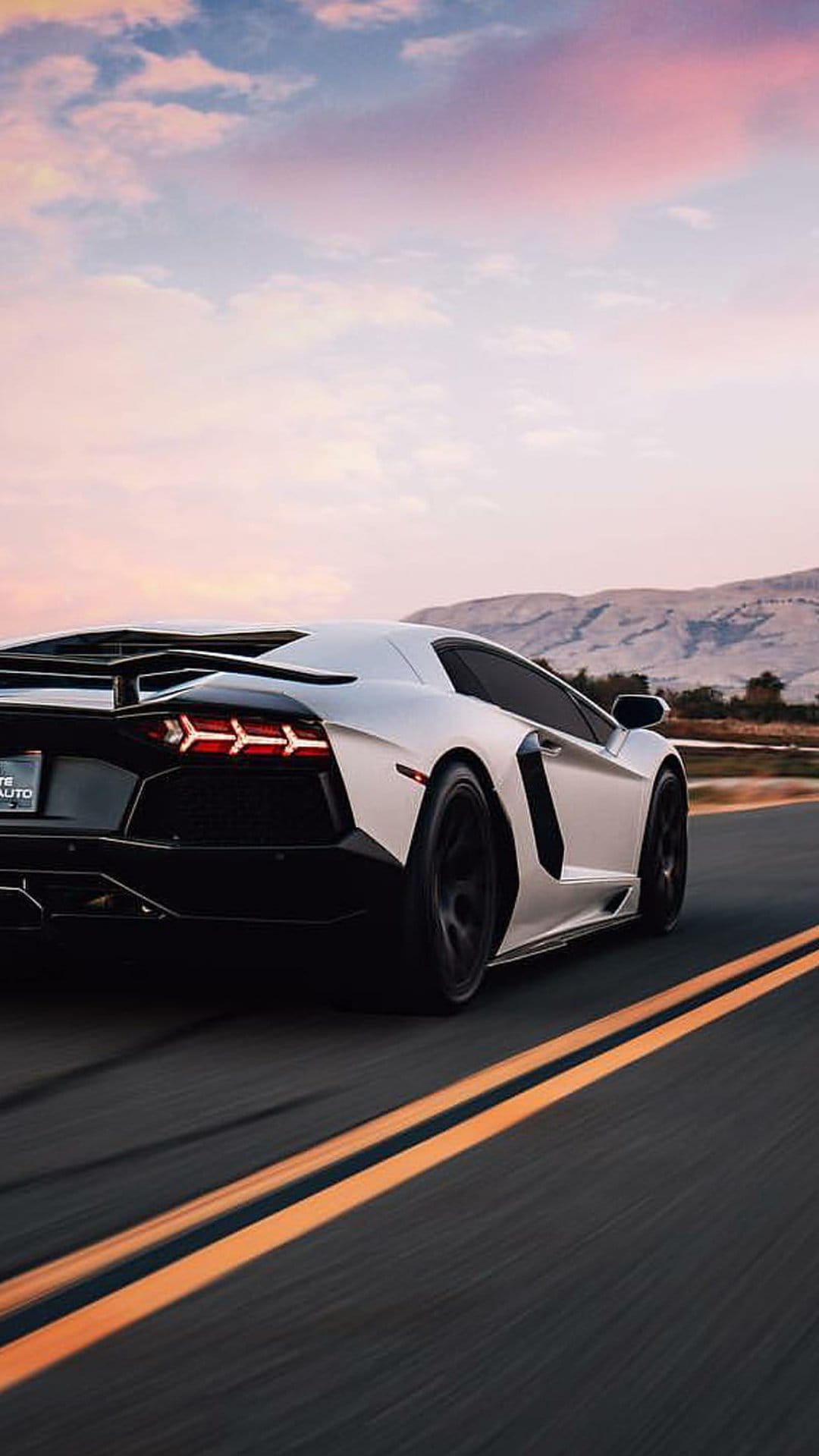 White Lamborghini Aventador with glowing taillights driving on a mountain road during a vibrant orange sunset.