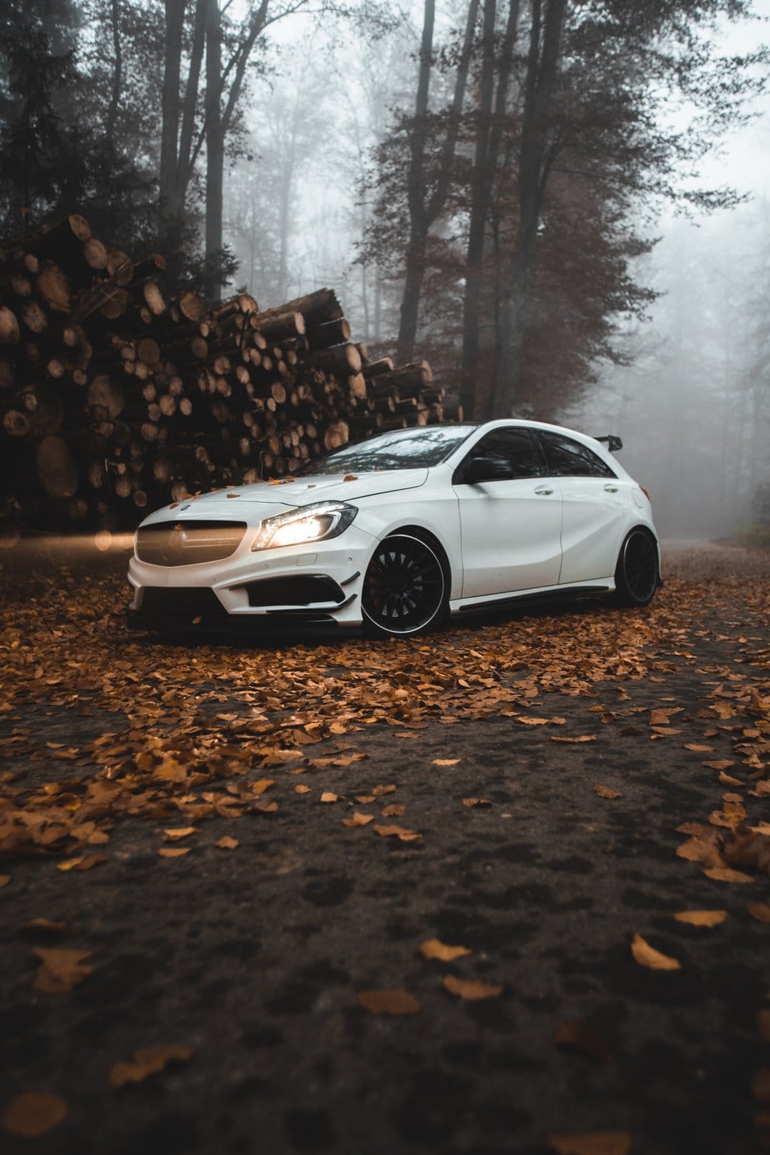 White Mercedes hatchback with headlights on parked on a misty forest road covered in orange autumn leaves.