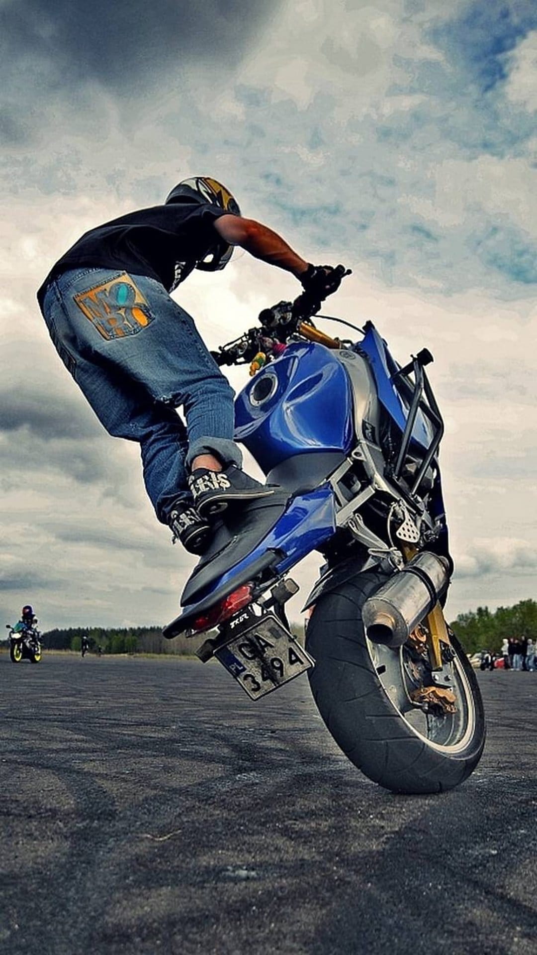 Motorcyclist in a helmet and jeans performs a wheelie on a blue sportbike under a cloudy sky on open asphalt.