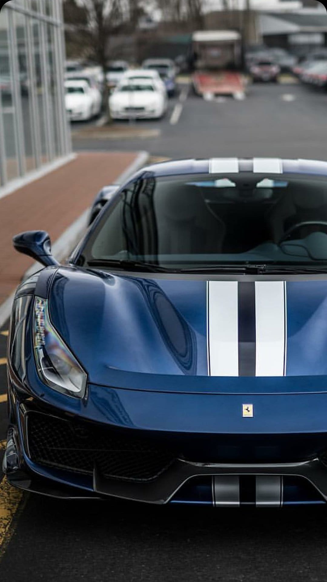 Blue Ferrari with white racing stripes on the front hood parked in an outdoor lot with a blurred building behind.