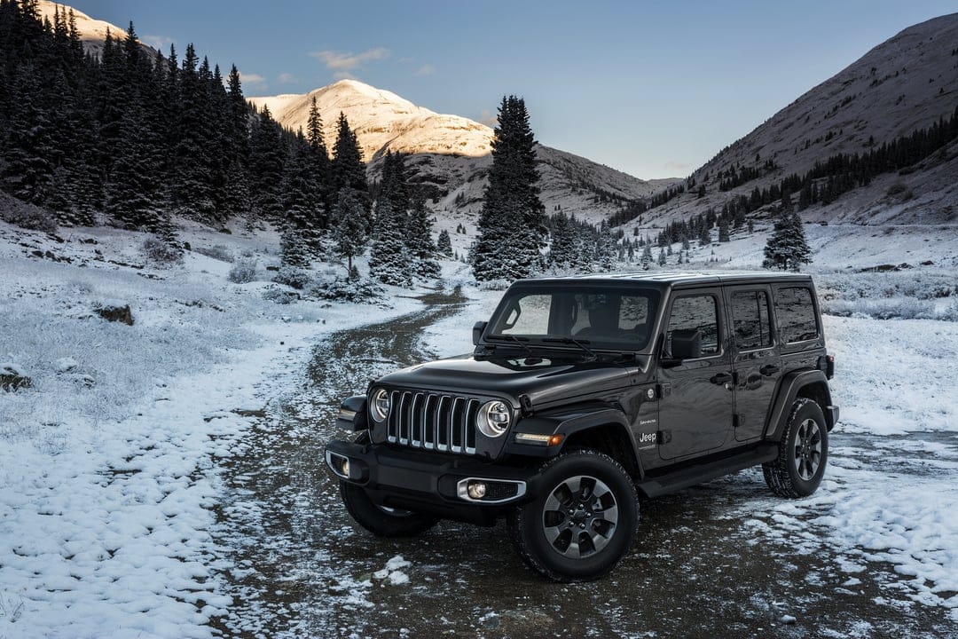 Black Jeep Wrangler Sahara parked on a dirt road in a snowy mountain valley with pine trees and peaks.