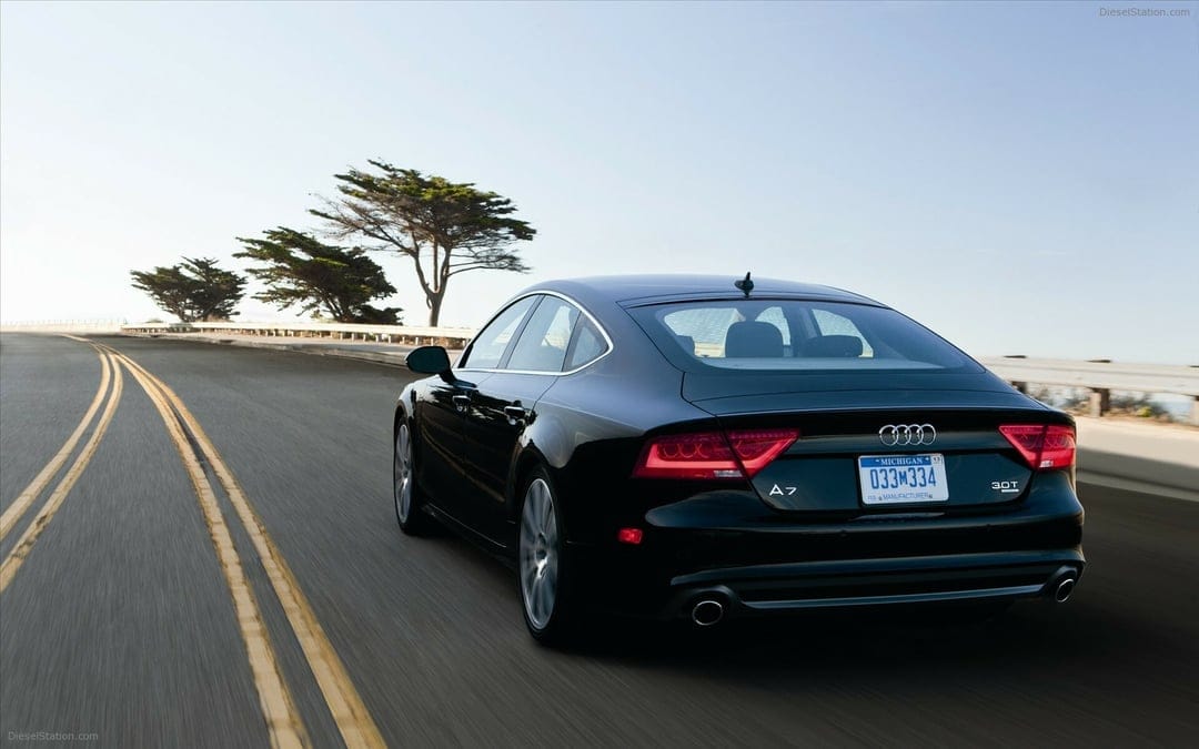 Black Audi A7 3.0T driving on a curved road with motion blur, coastal trees, and clear sky in the background.