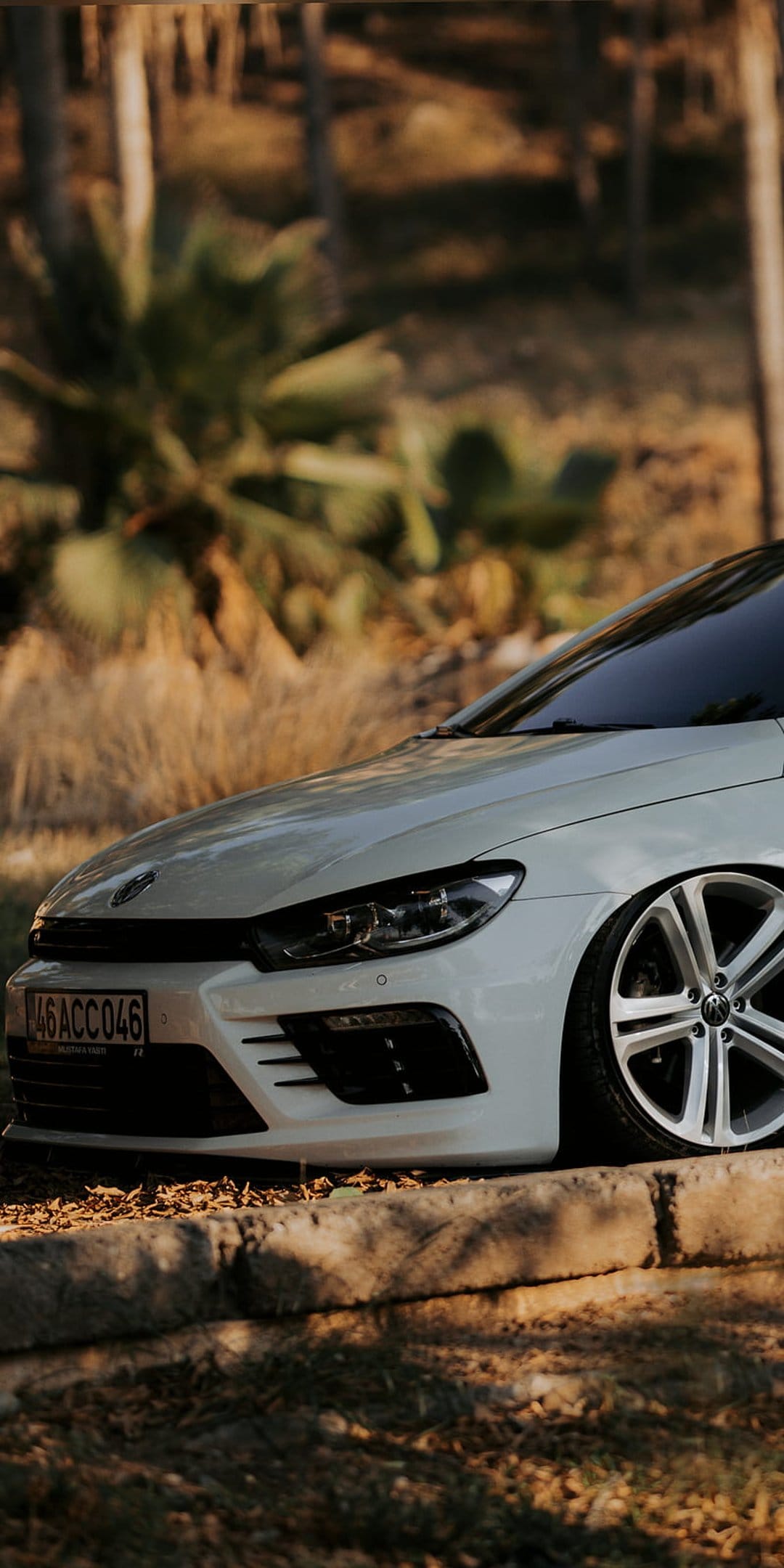 White Volkswagen Scirocco with custom wheels and sharp headlights parked on grass in warm sunset lighting.