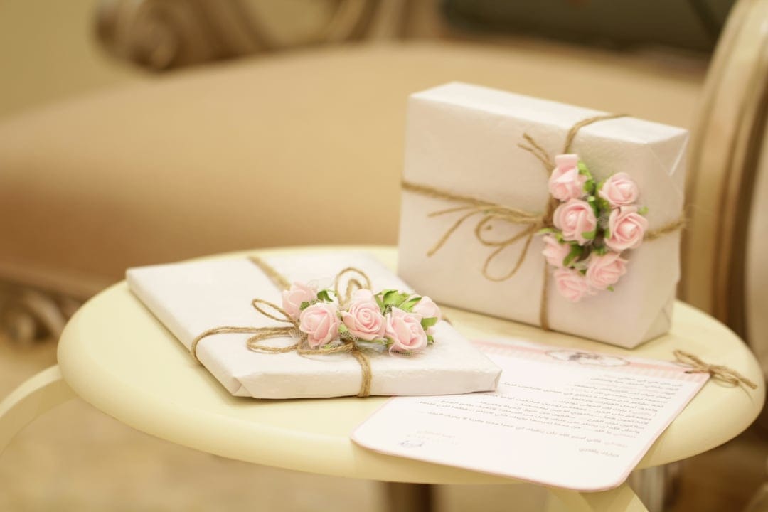 White wrapped gift boxes with pink roses and rustic twine on a yellow table with a handwritten card.