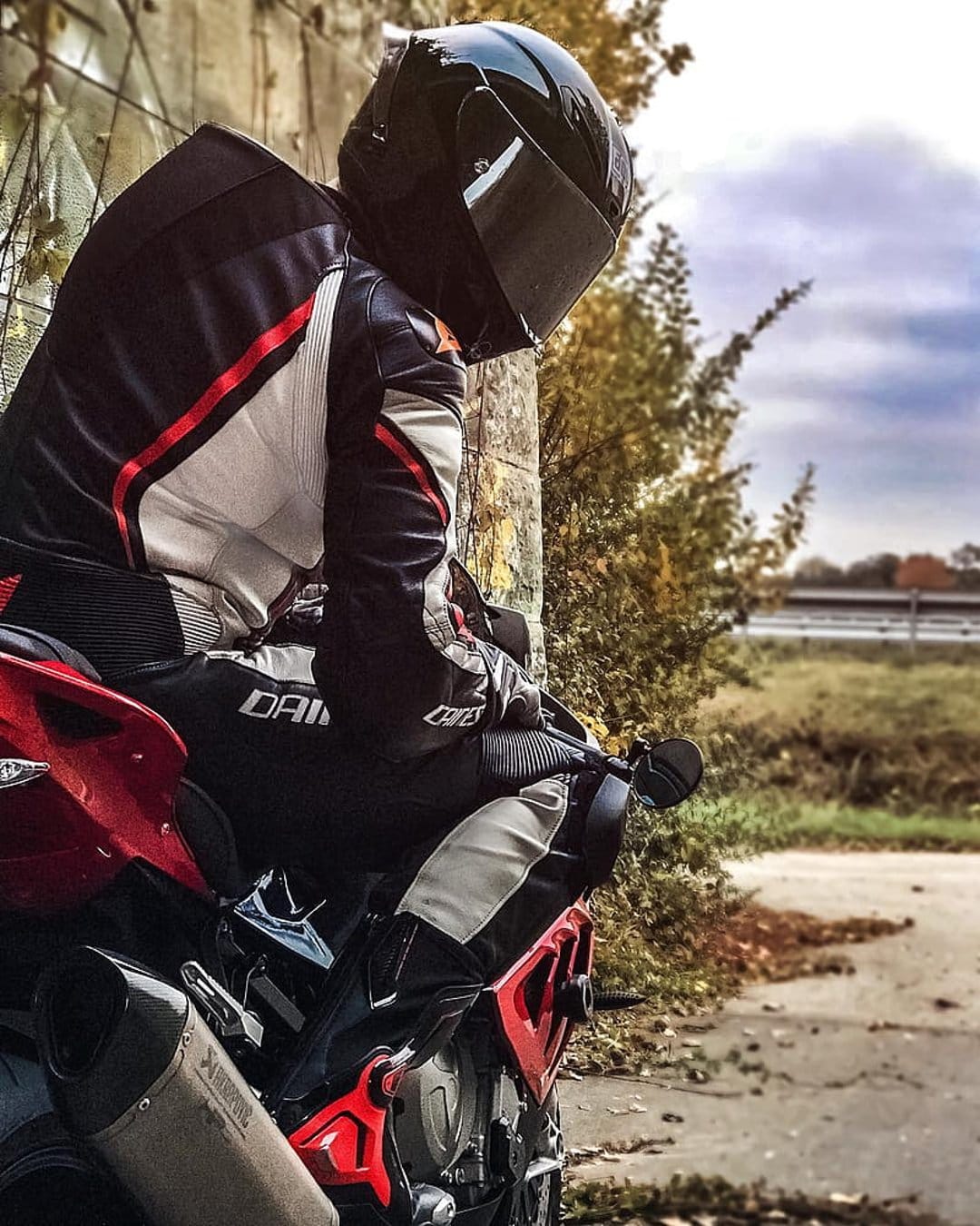 Motorcyclist in black and white leathers sits on a red bike against a concrete wall under a cloudy autumn sky.