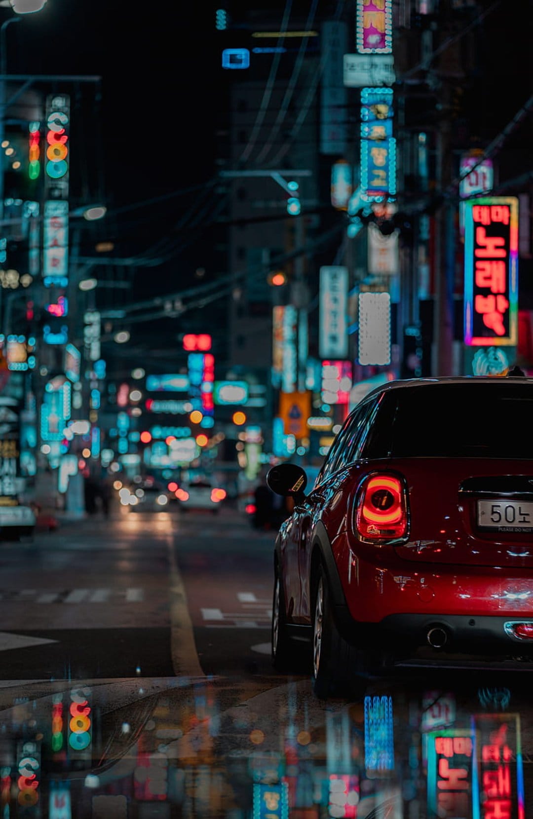 Red Mini Cooper with glowing taillights parked on a wet street under vibrant Korean neon signs at night.