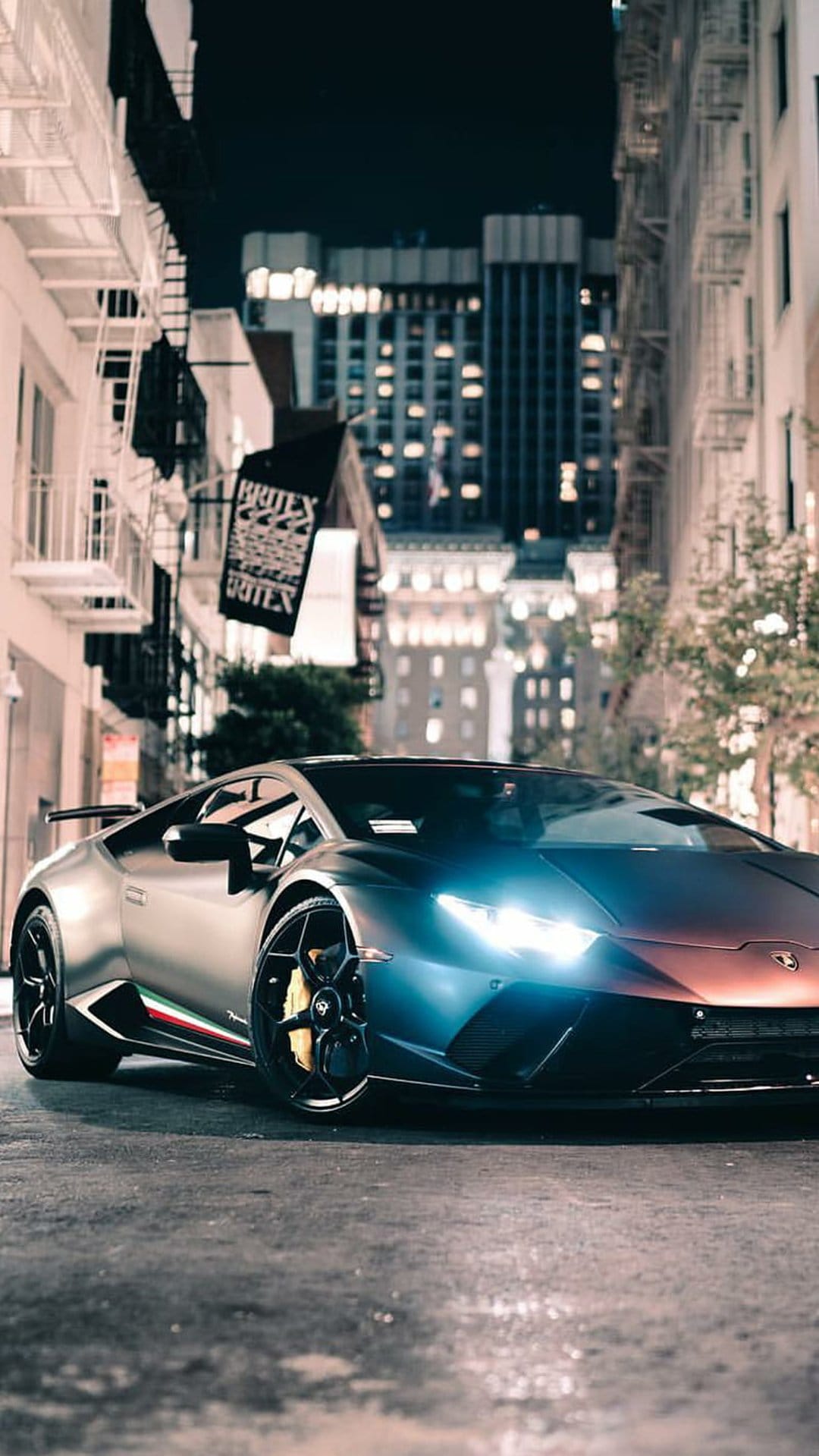 Lamborghini supercar with glowing headlights parked on wet city pavement under glowing urban building lights.