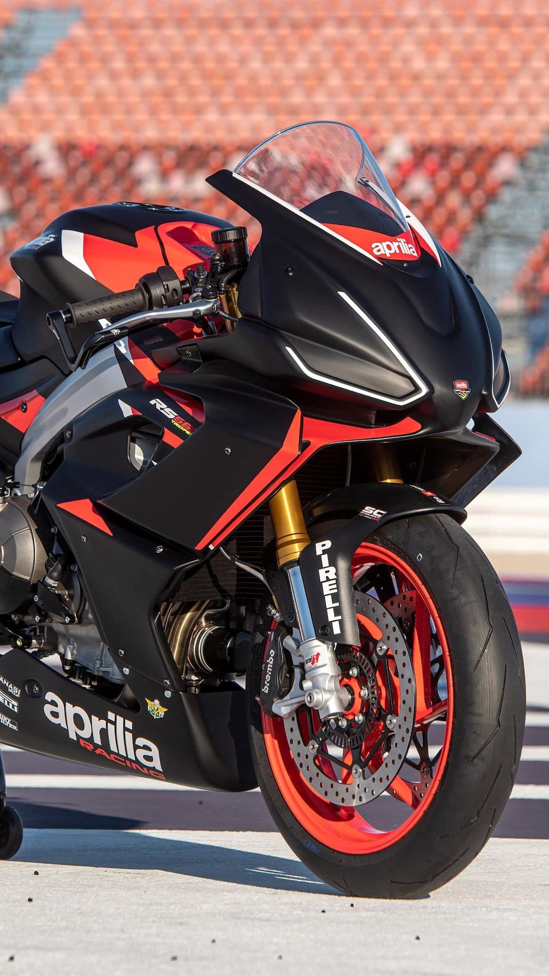 Close-up of a black and red Aprilia racing motorcycle with gold forks and red wheels on a sunny track.