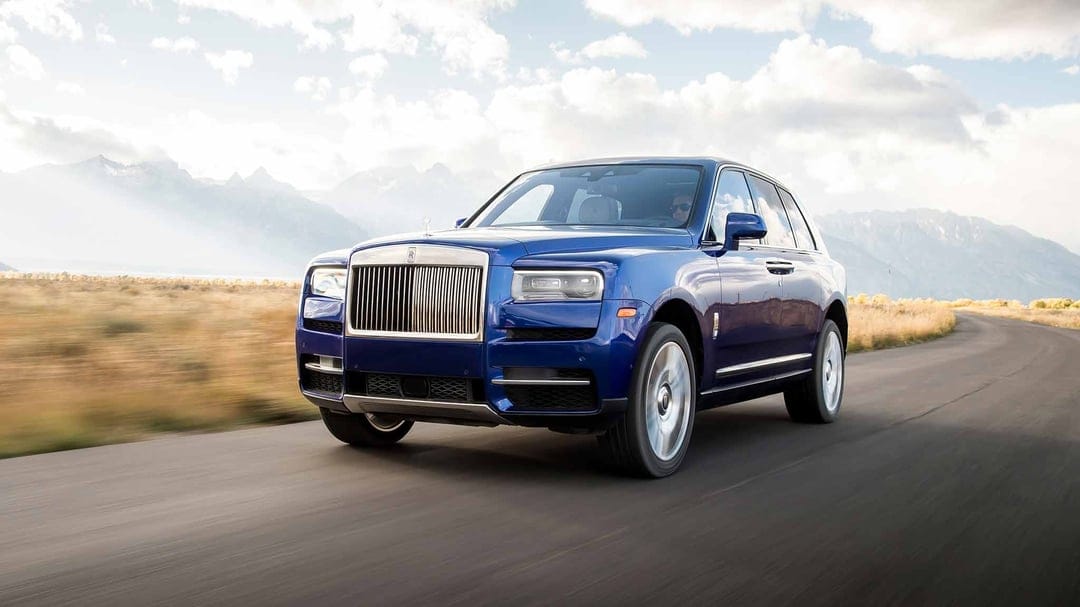 Blue Rolls-Royce Cullinan driving on an open road with misty mountains and a cloudy sky in the background.