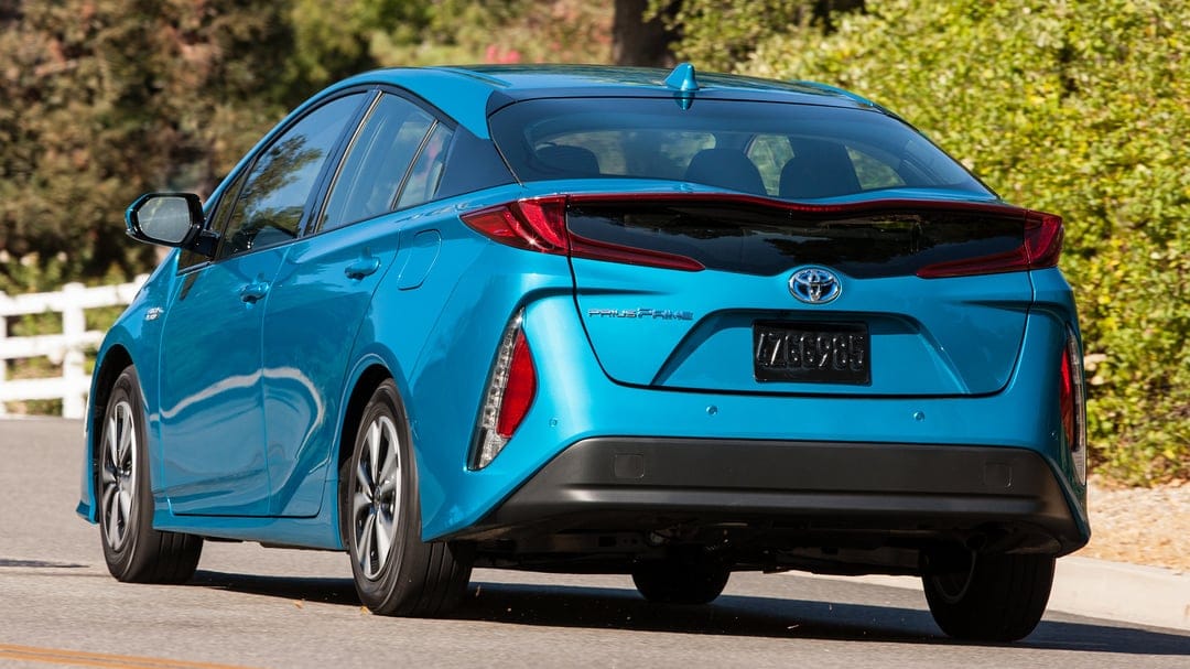 Rear view of a metallic blue Toyota Prius Prime driving on an asphalt road past green trees and a white fence.