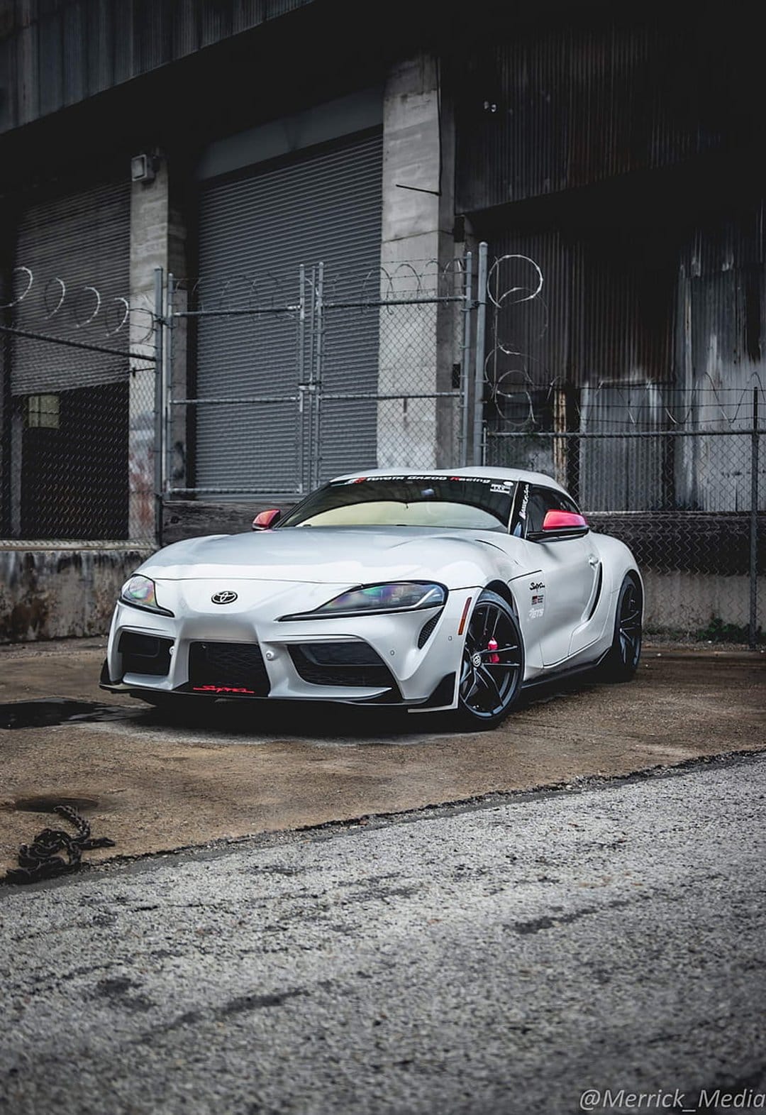 White Toyota Supra sports car with red accents parked near a chain-link fence and weathered industrial building.