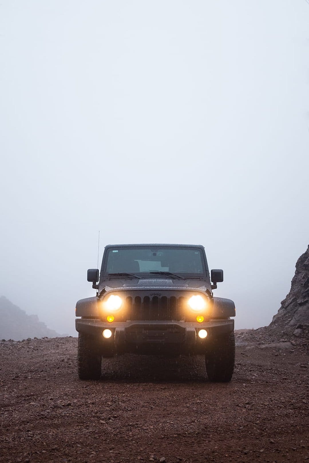 Black Jeep Wrangler with glowing headlights driving on a rocky mountain road through thick, atmospheric gray fog.