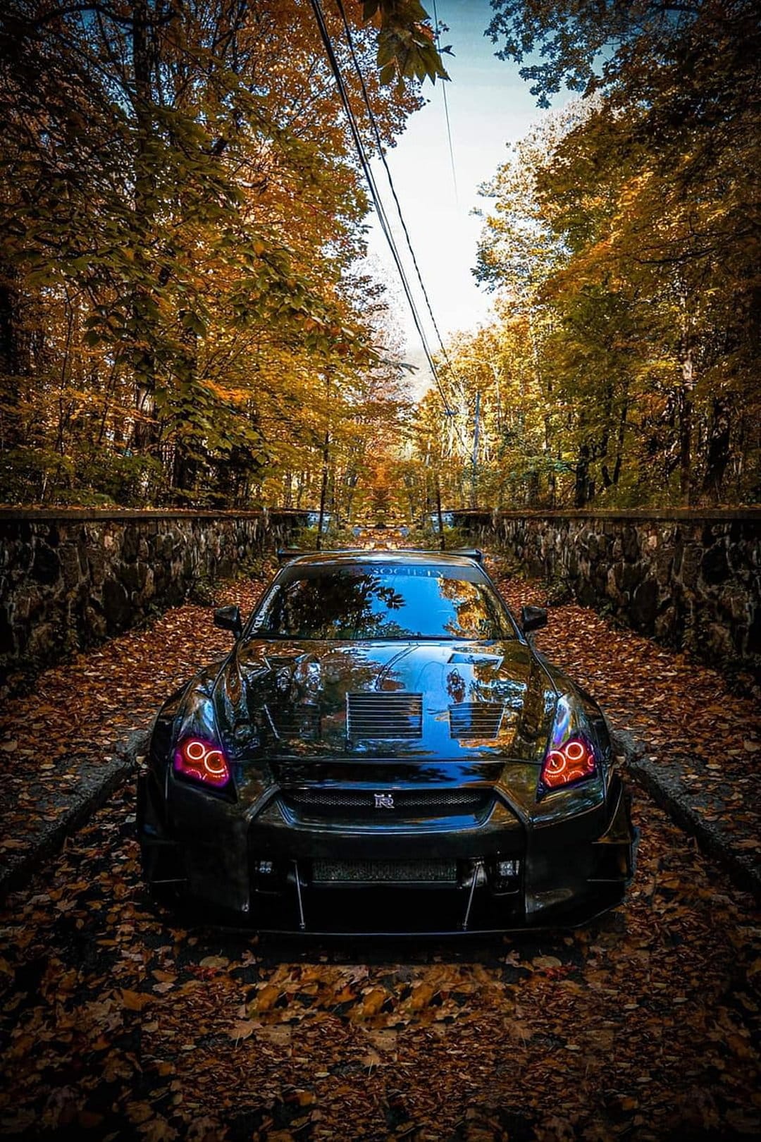 Black Nissan GT-R sports car with red halo headlights on a leaf-strewn road surrounded by orange autumn trees.