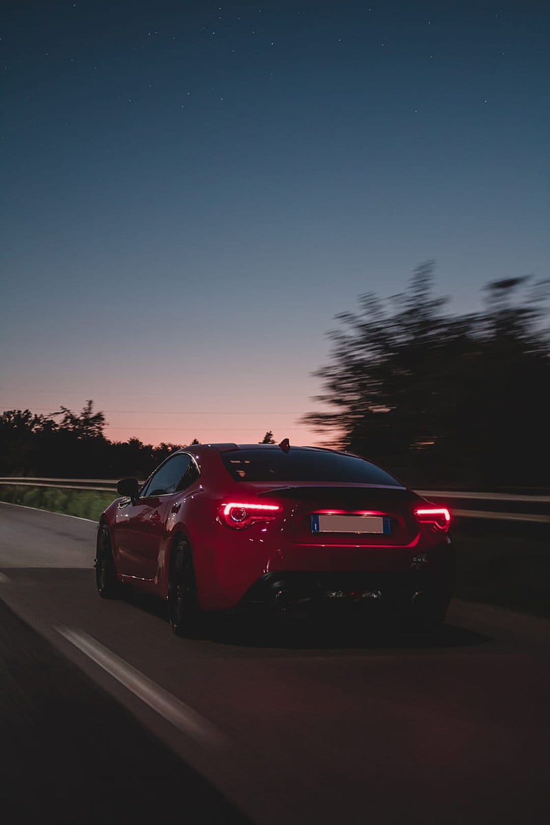 Red sports car driving on a road at night with glowing taillights under a starry sky and orange horizon.