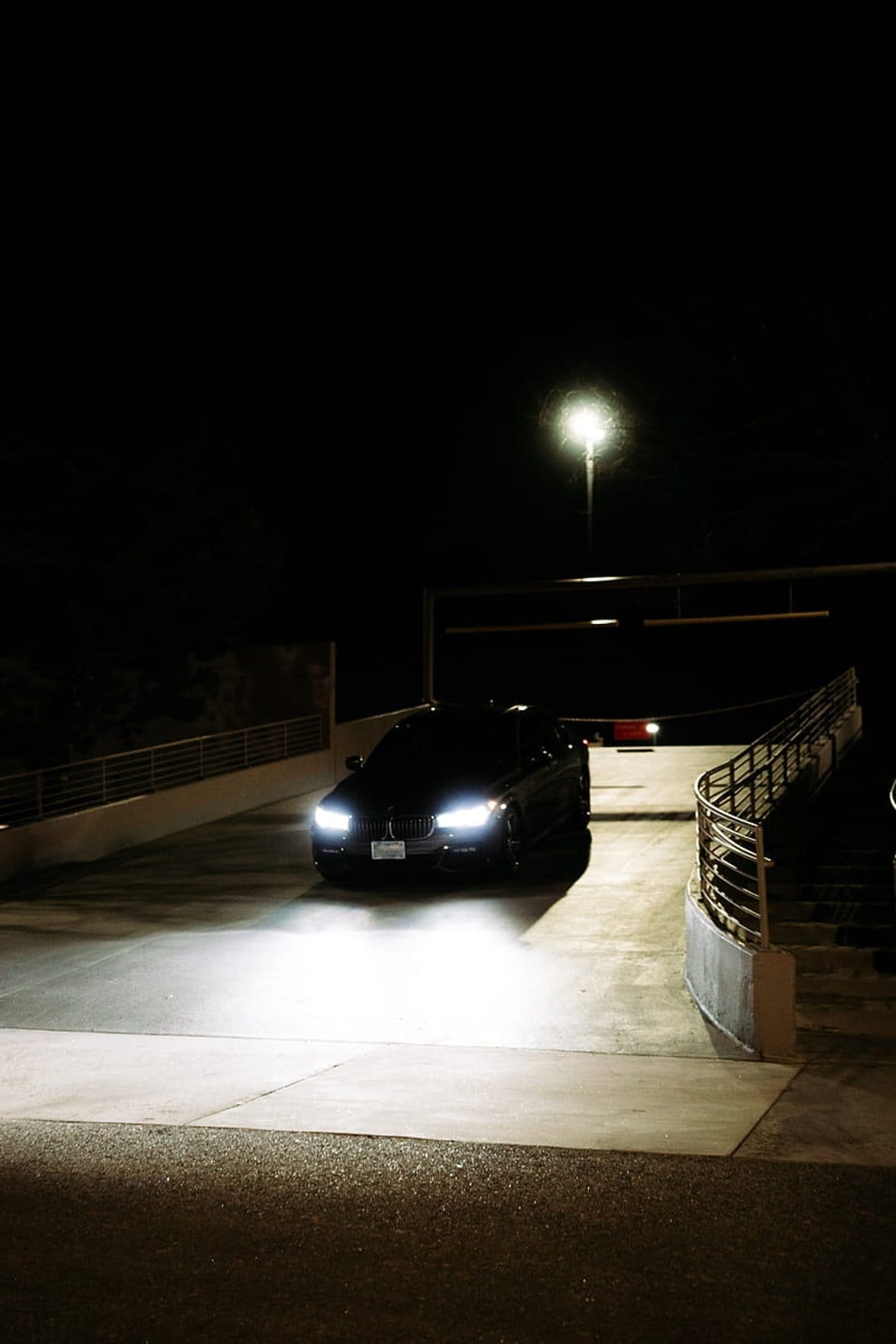 Black BMW sedan drives through a dark concrete parking garage with bright headlights and an overhead streetlamp.