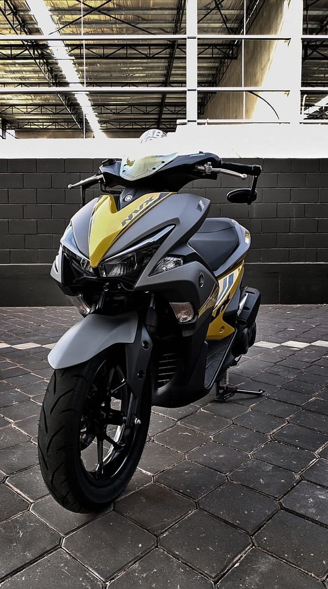 Yellow Yamaha NVX scooter parked by a brick wall with gray accents and sharp aerodynamic fairings on paved ground.
