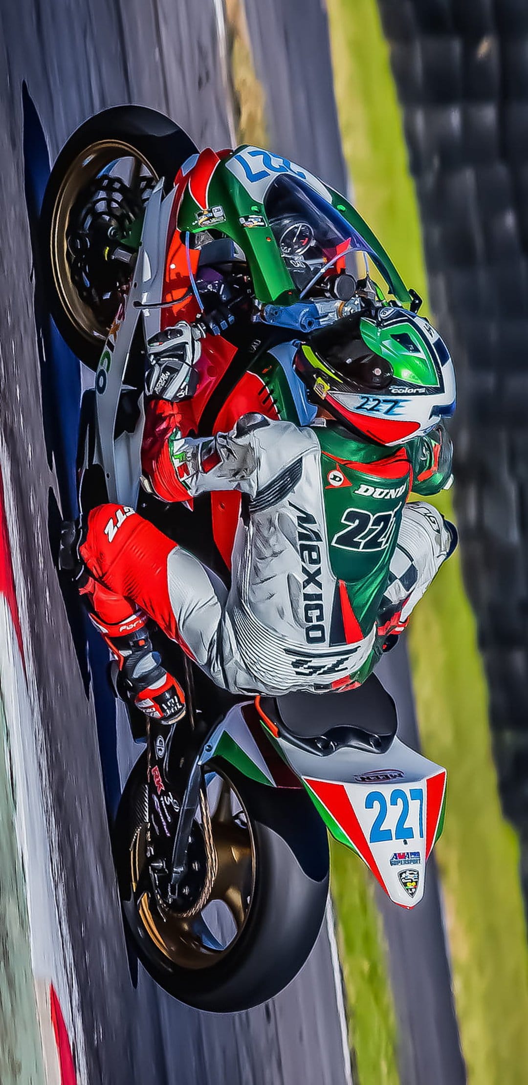 Professional racer in green and white gear leans a number 227 sportbike deep into a racetrack curve at high speed.