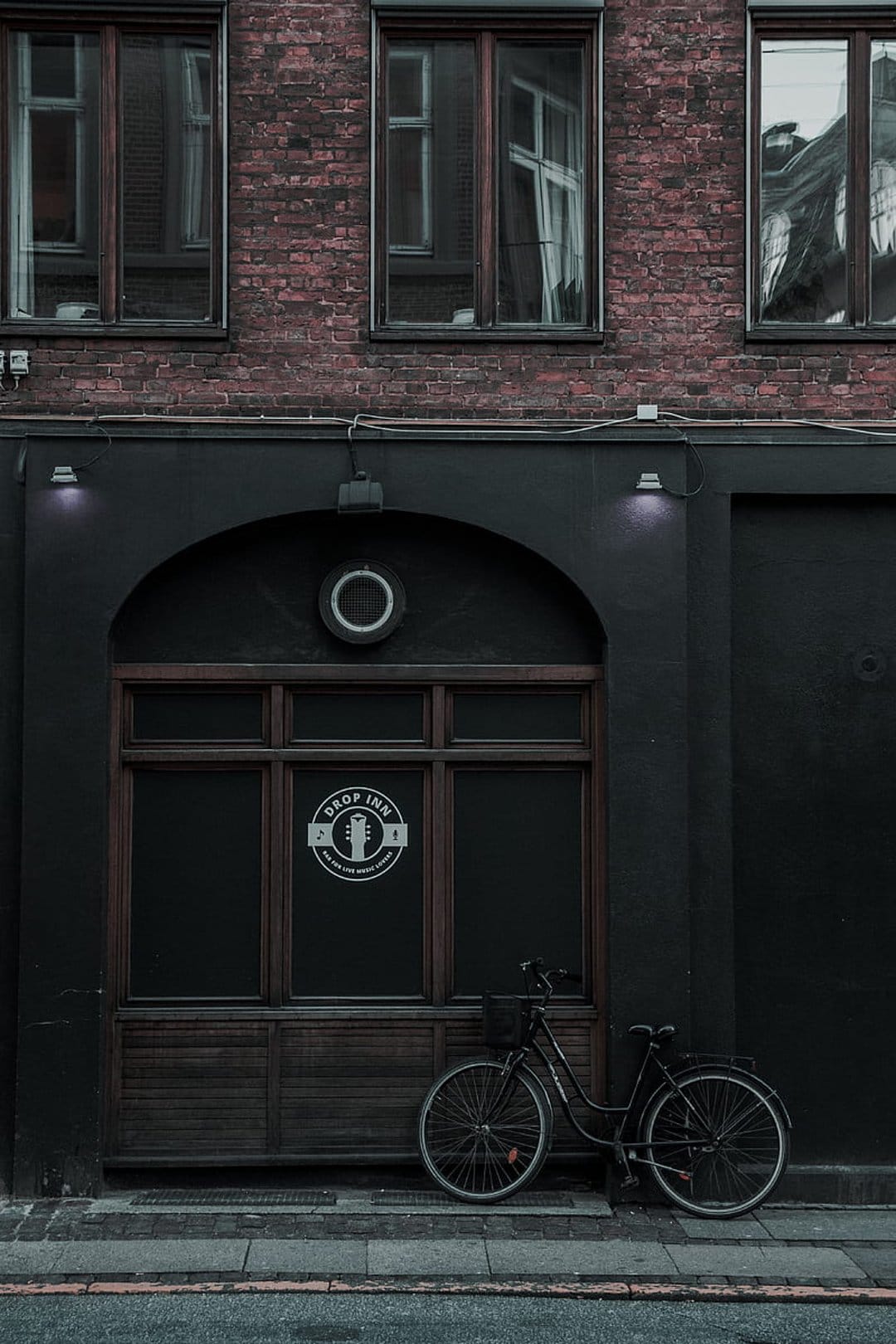 Vintage bicycle leans against a dark bar entrance below a red brick building during a moody urban evening.