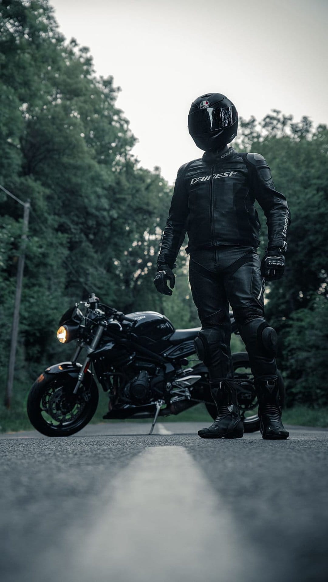 Biker in black gear stands next to a sportbike with its headlight on against a backdrop of dense green trees.