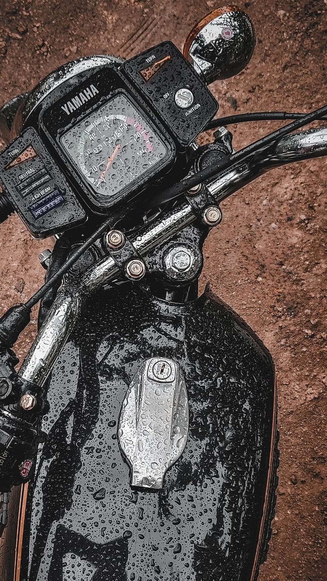 Yamaha motorcycle handlebars and chrome fuel cap covered in water droplets with a blurred outdoor background.