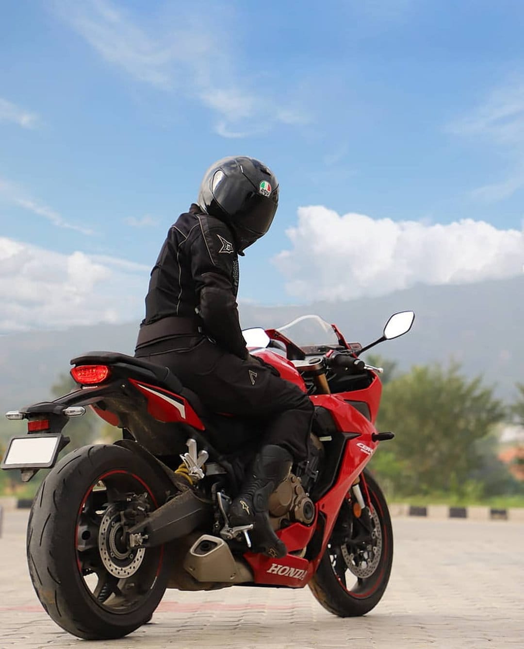 Motorcyclist in black gear sits on a red sportbike against a backdrop of blue sky, white clouds, and green hills.