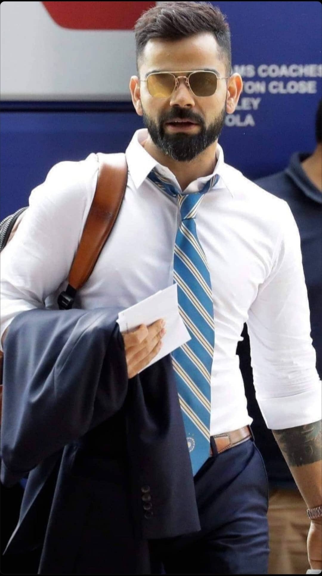 Virat Kohli in a white shirt and striped tie wears aviator sunglasses and carries a dark blazer and brown bag.