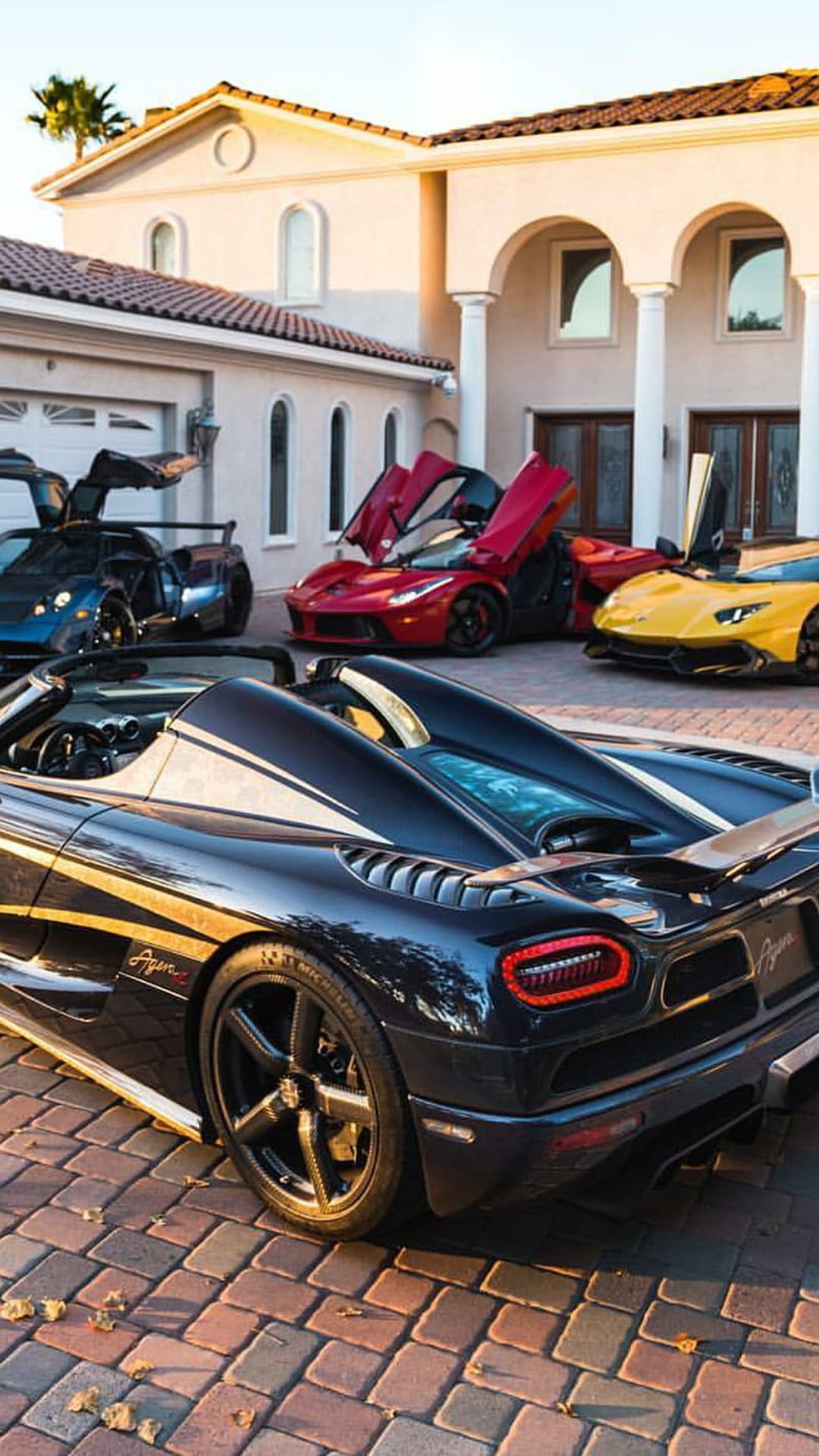 Multi-colored luxury hypercars with open doors parked on a stone driveway in front of a grand mansion facade.