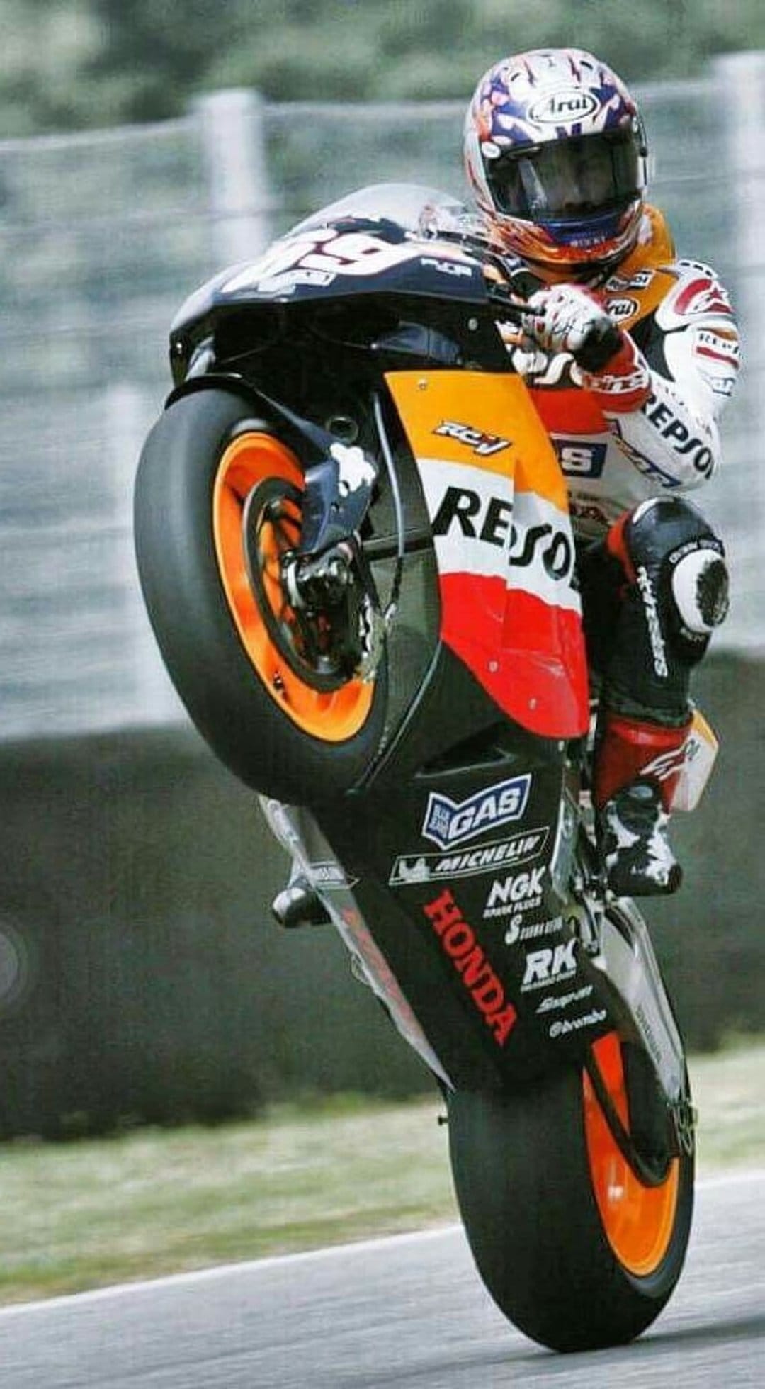 Professional motorcycle racer in an orange suit performs a wheelie on a blurred asphalt race track.