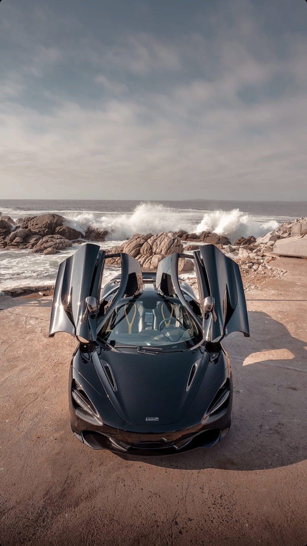 Black McLaren supercar with open doors parked on a sandy beach with crashing ocean waves and coastal rocks.