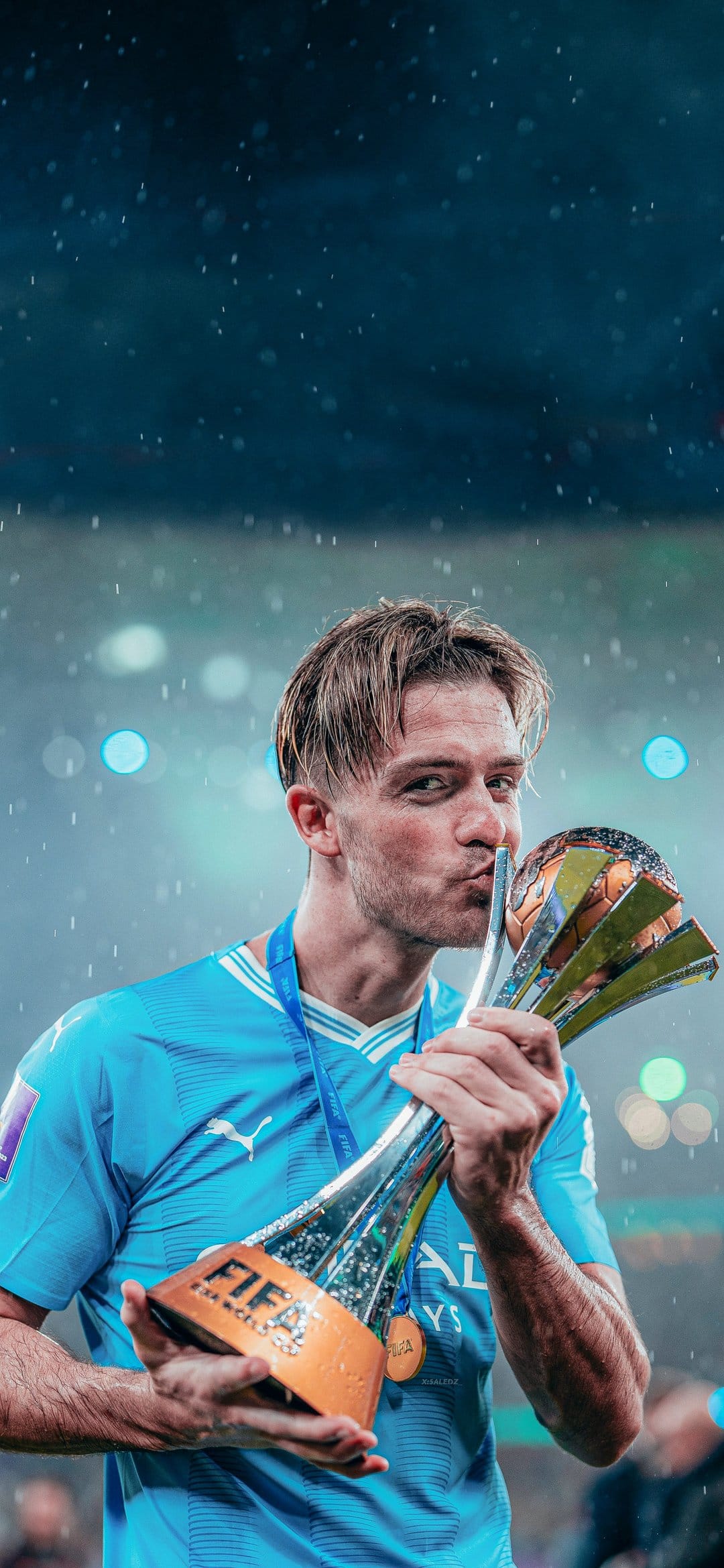 Jack Grealish kissing the gold FIFA Club World Cup trophy in the rain while wearing a sky blue jersey and medal.