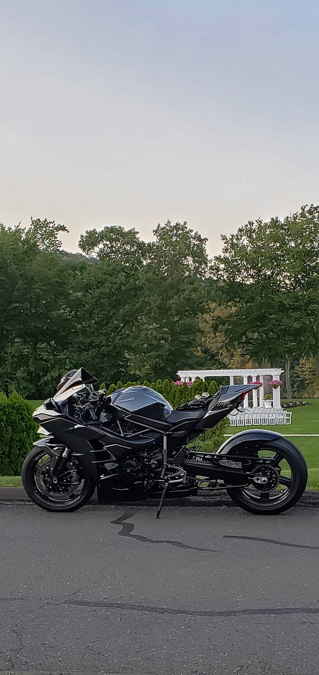Black Kawasaki motorcycle parked on asphalt with green trees and a white building under soft morning sunlight.