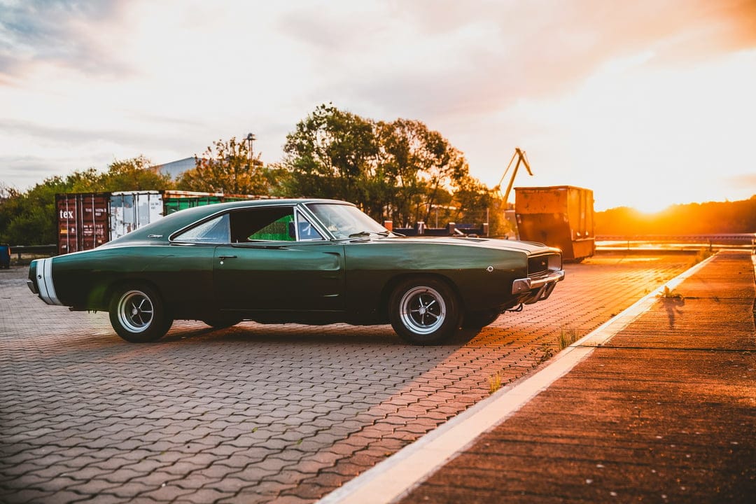 Green Dodge Charger parked at an industrial shipyard during a vibrant orange sunset with cranes in the background.