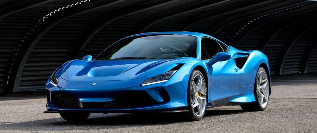 Electric blue Ferrari F8 Tributo with silver wheels parked under a dark ribbed architectural structure on asphalt.