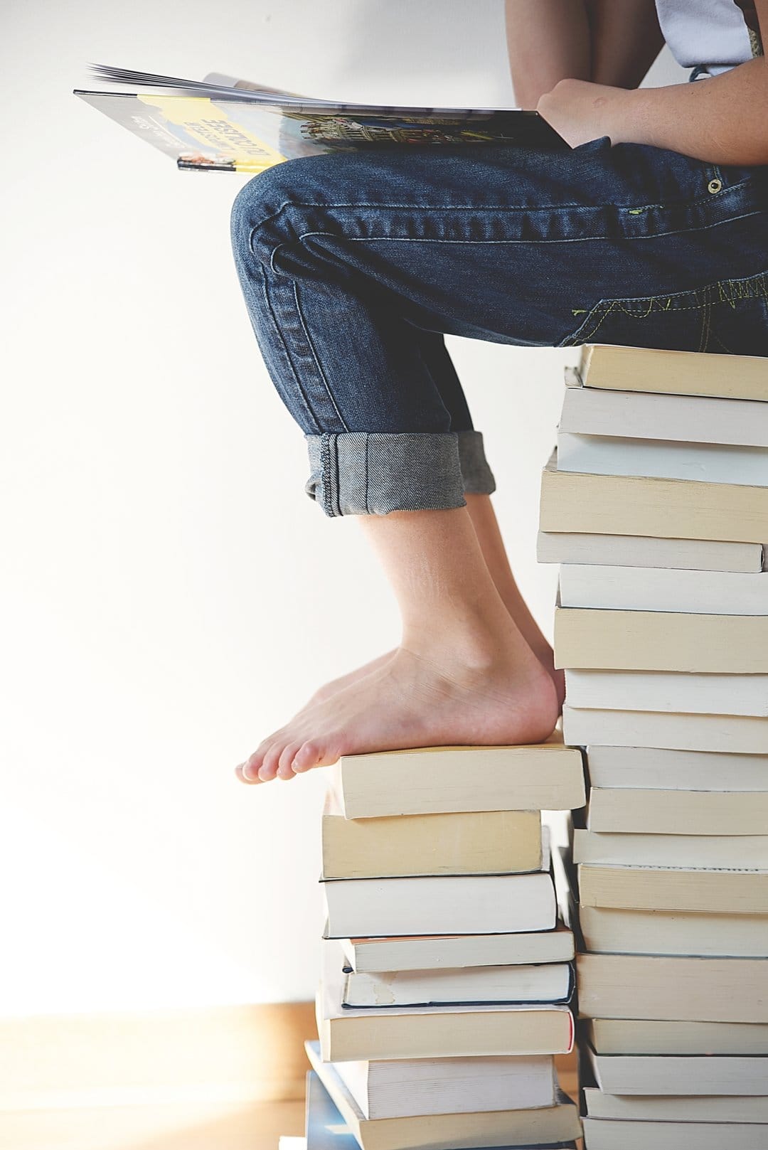 Young person in cuffed denim jeans sits atop a tall stack of books reading against a solid white background.