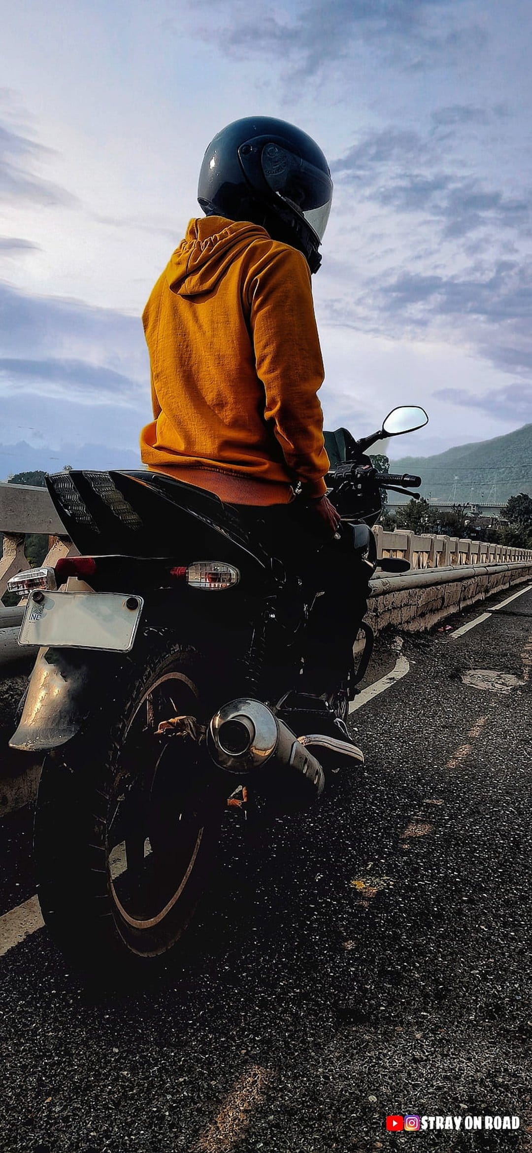 Motorcyclist in an orange hoodie on a bridge overlooking a winding road through lush green hills under a cloudy sky.