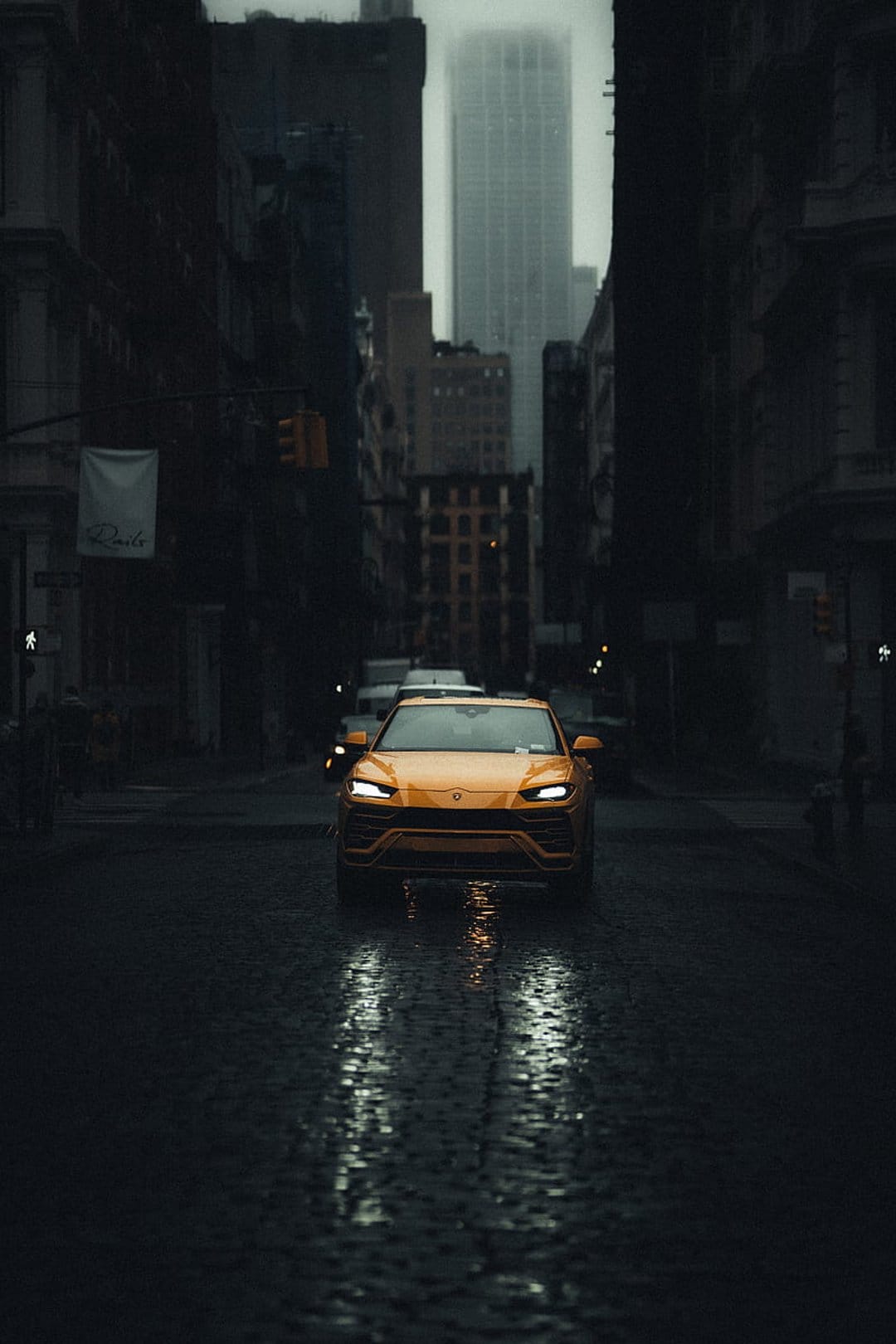 Yellow Lamborghini Urus driving on a wet cobblestone street at dusk with glowing headlight reflections.