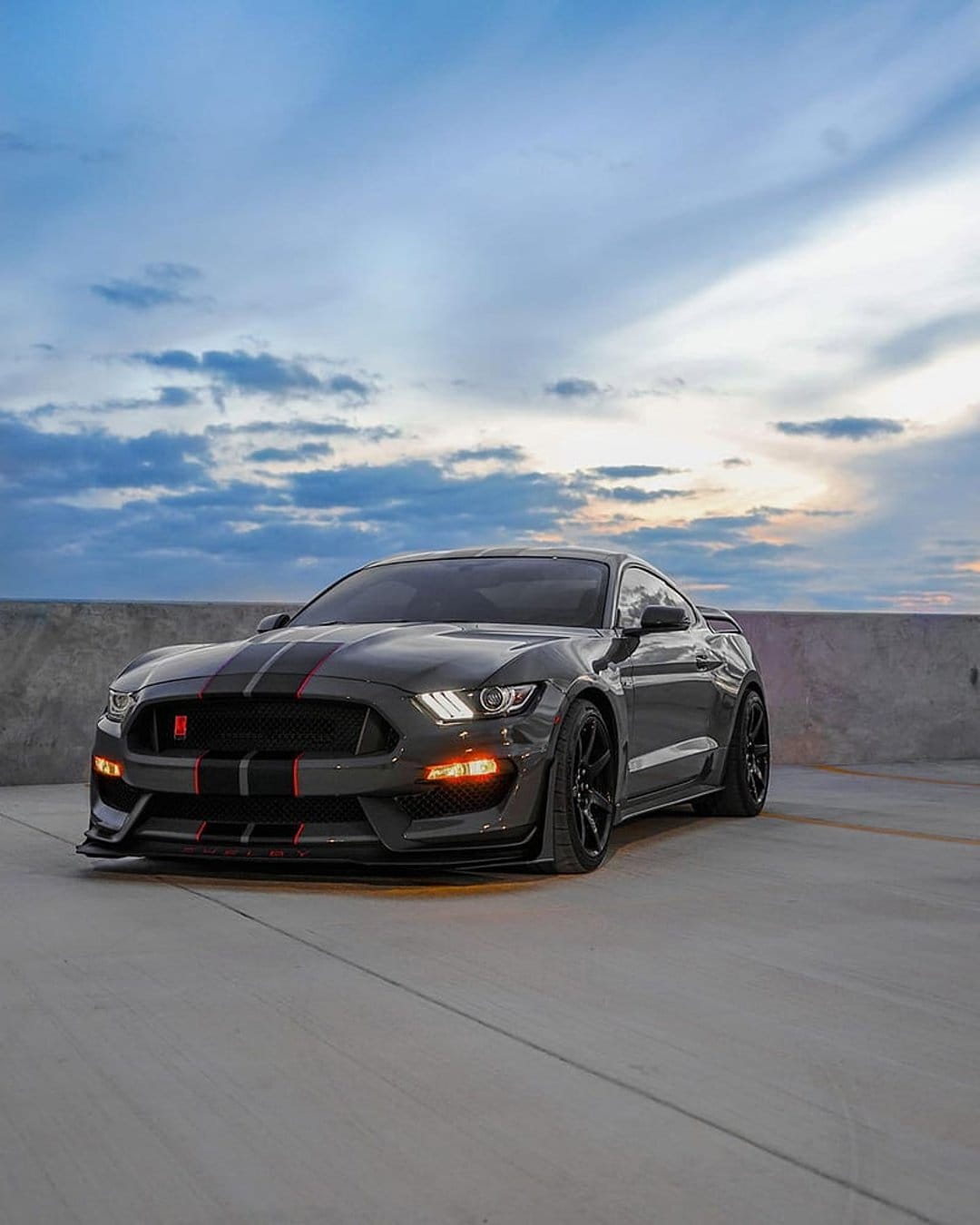 Ford Mustang GT350 with red stripes parked on concrete under a blue and white twilight sky in portrait view.
