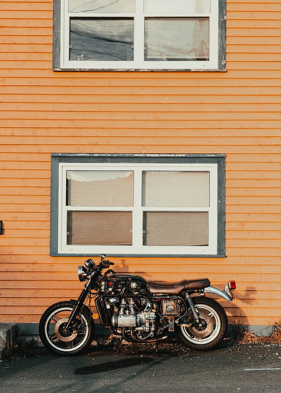 Vintage black motorcycle with a brown leather seat parked against bright orange house siding with white windows.