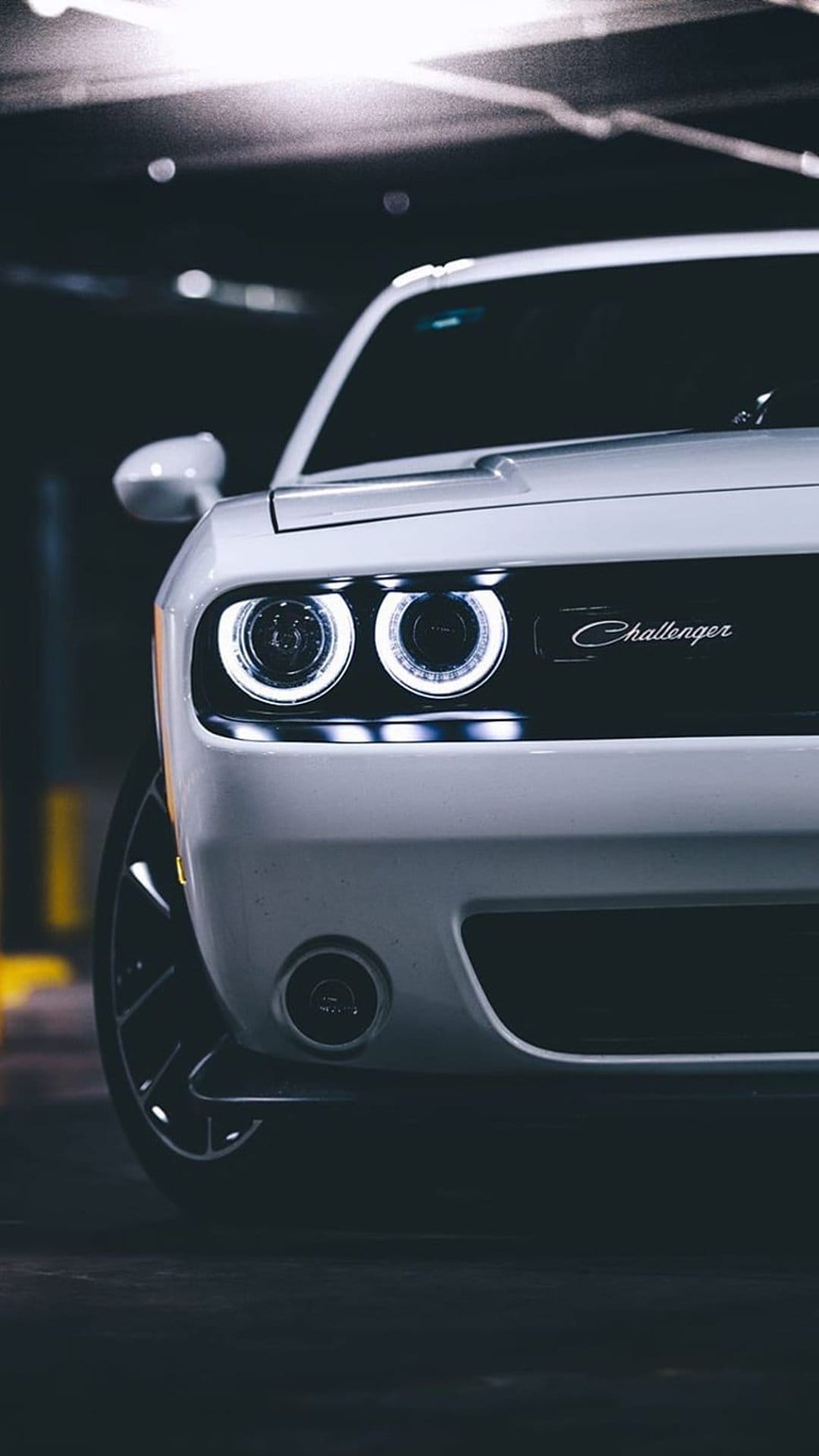 White Dodge Challenger front grille and glowing circular LED headlights in a dark parking structure with bokeh.