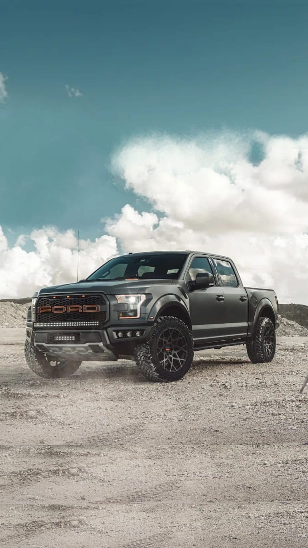 Ford F-150 Raptor pickup with orange lettering and LED lights parked on a rocky desert trail under a blue sky.
