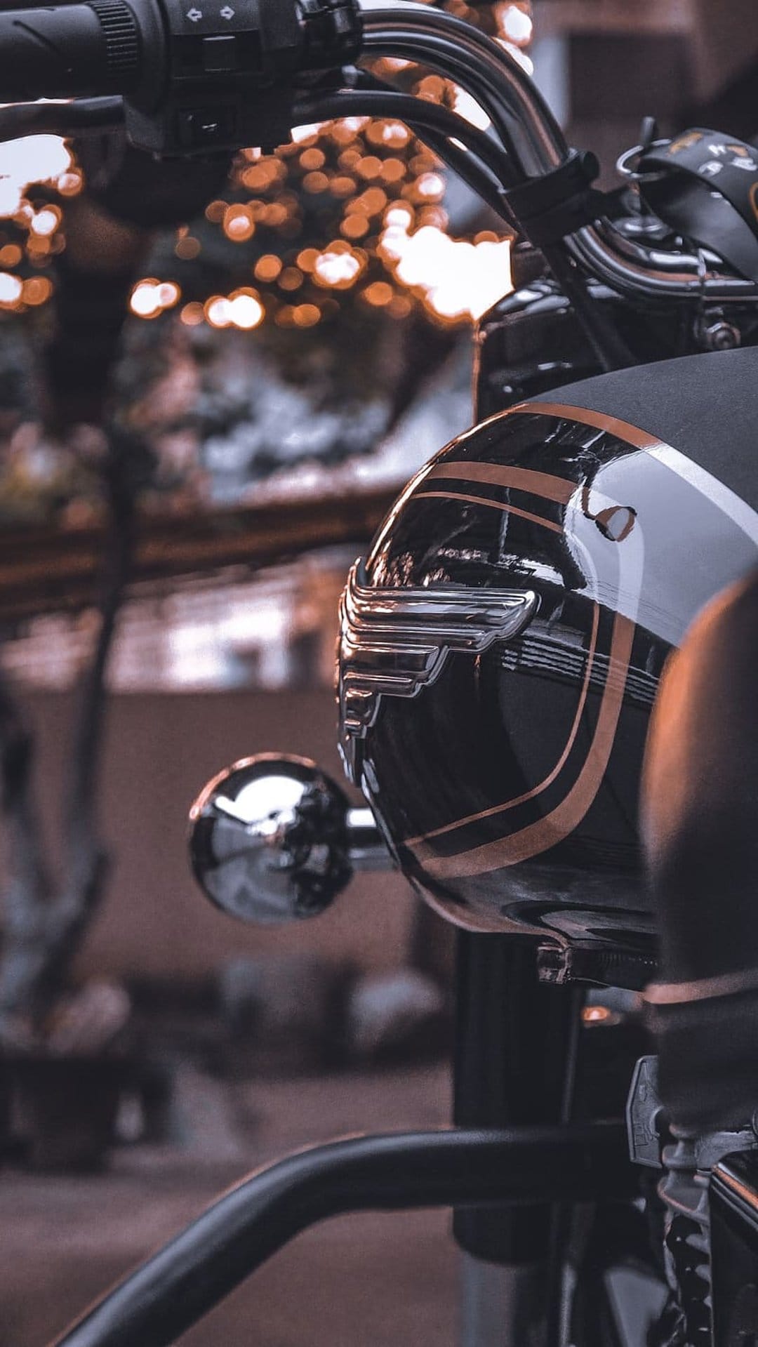 Polished black vintage motorcycle headlight with a chrome winged emblem and gold stripes against orange bokeh.
