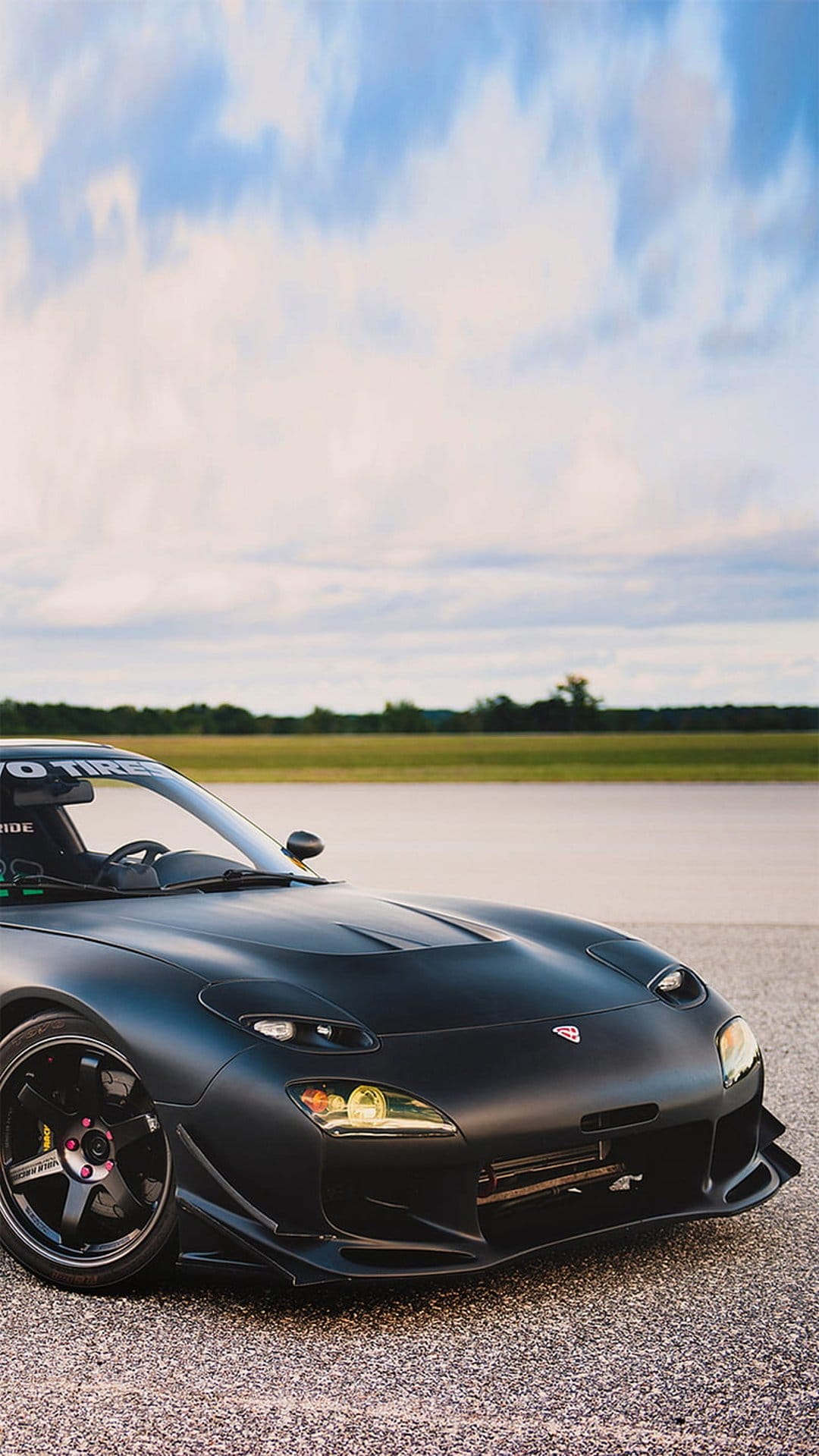Mazda RX-7 in matte black parked on gravel with an aggressive body kit under a moody blue and white cloudy sky.
