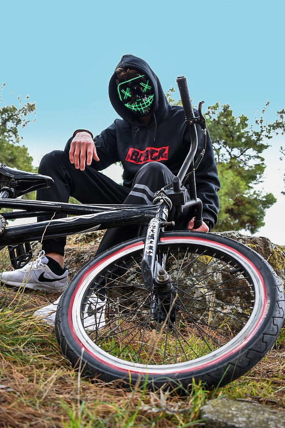 Person in a black hoodie and glowing neon green mask sits on a black BMX bike against a grassy forest backdrop.