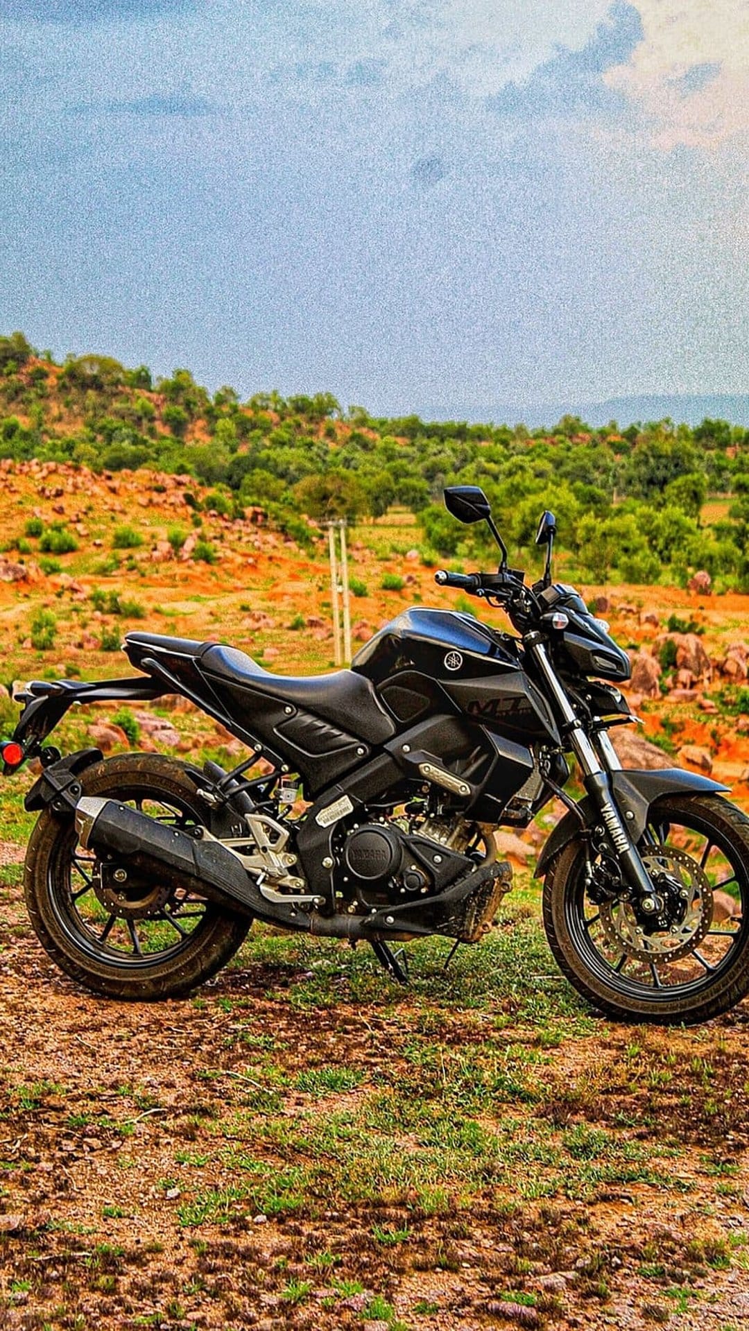 Black Yamaha MT-15 motorcycle parked on a dirt trail surrounded by green foliage under a cloudy grey sky.