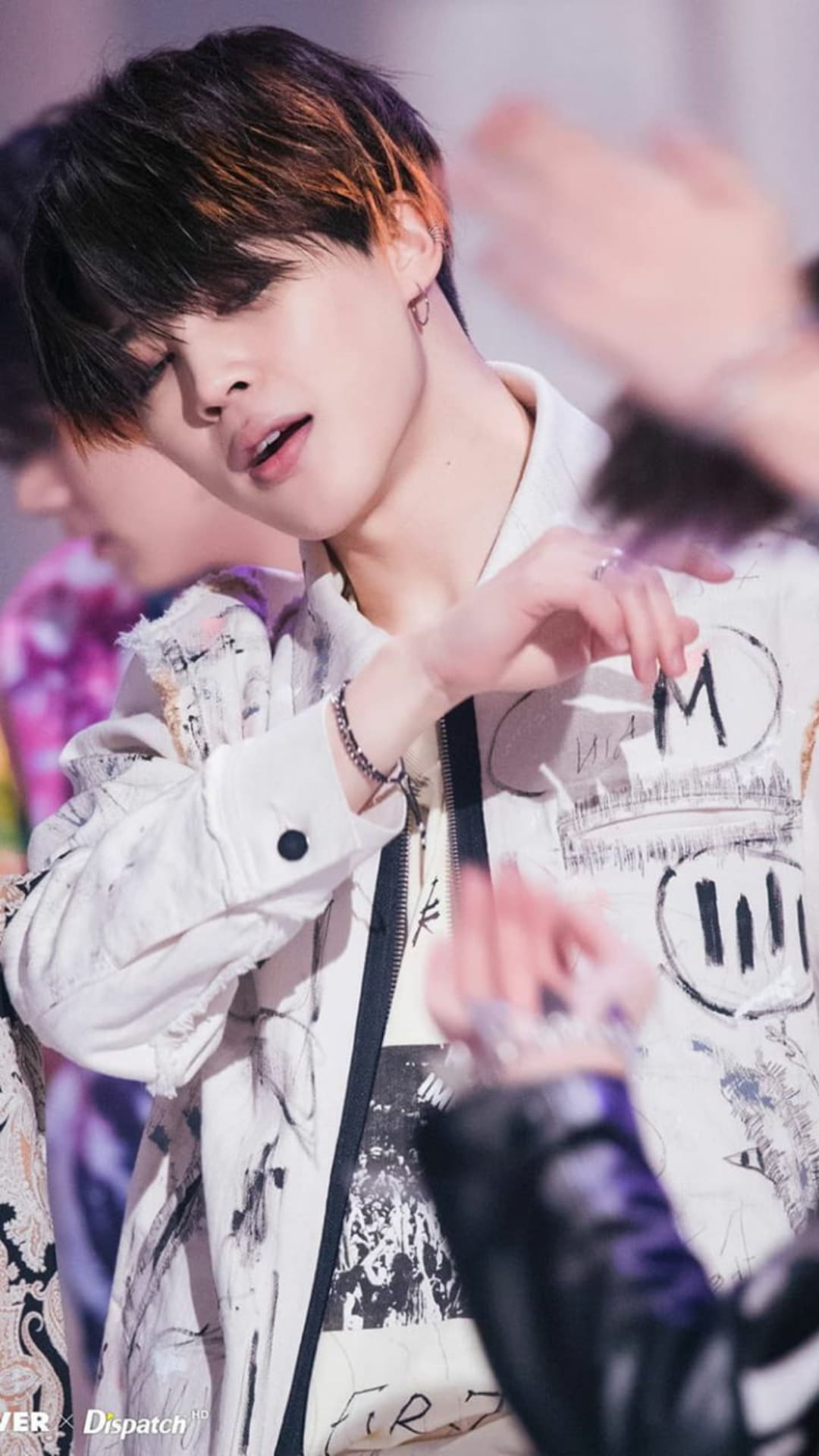 Park Jimin of BTS tilts his head down wearing a white graffiti-print jacket and silver rings during a concert.