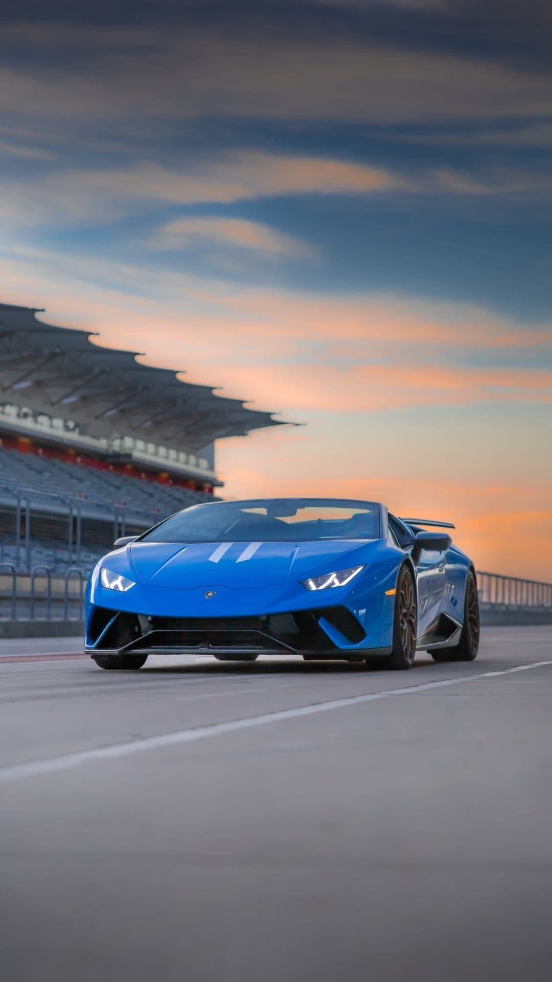 Blue Lamborghini convertible on a race track under a sunset sky with a stadium grandstand in the background.