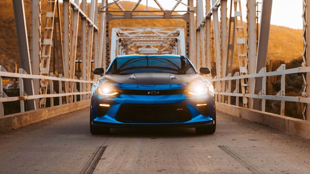 Front view of a blue Chevrolet Camaro SS with black hood on a steel bridge at sunset with glowing headlights.