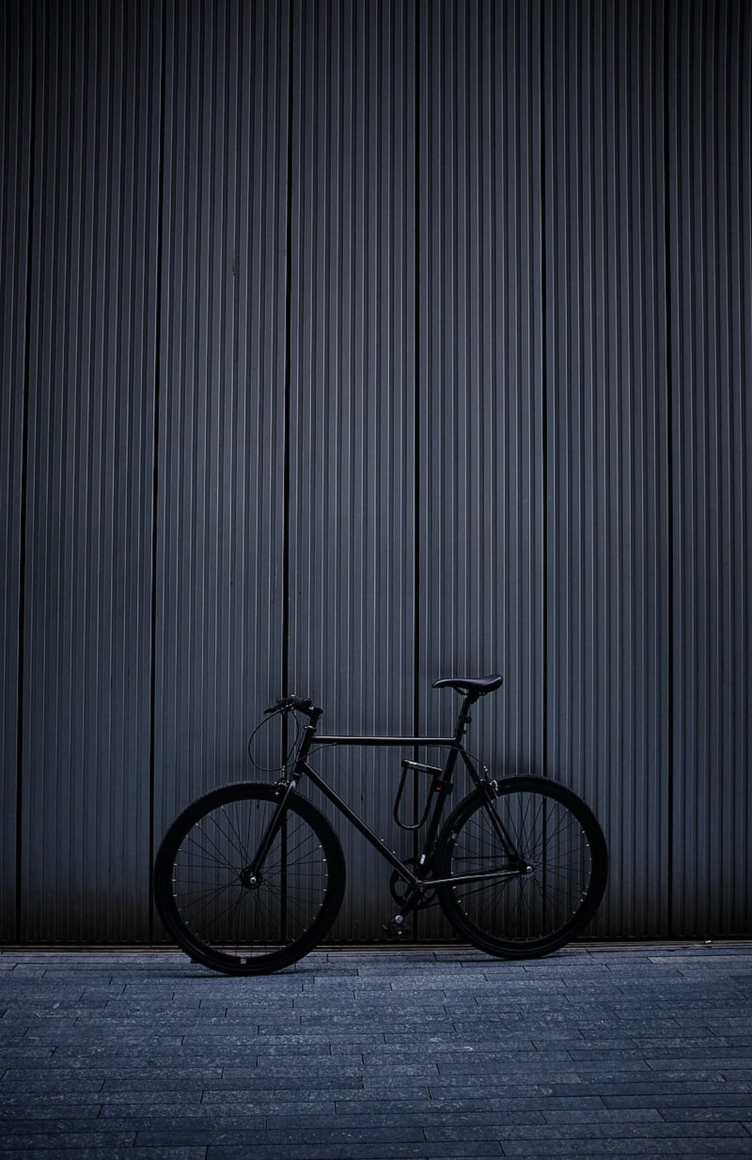 Black bicycle with thin tires parked against a vertically ribbed dark wall on a paved stone ground in soft light.