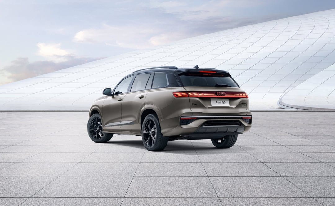 Rear view of a bronze Audi Q6 SUV on a gray tiled plaza with a curved white architectural background.