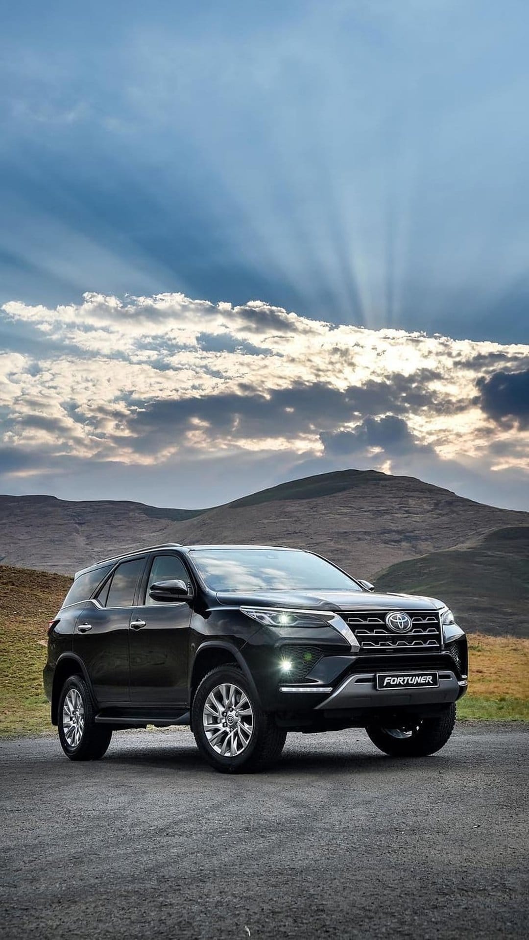 Toyota Fortuner SUV in black parked on a gravel mountain road with sun rays piercing through clouds in the sky.